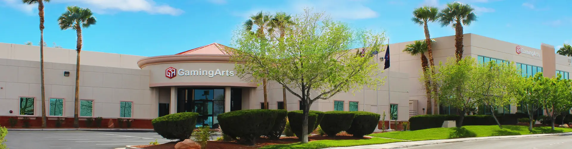A modern commercial building with a Gaming Arts sign, surrounded by trimmed bushes, green trees, and palm trees under a blue sky. Showcasing the latest in casino gaming technology, this impressive space welcomes visitors with its state-of-the-art features and convenient parking lot visible in front of the entrance.