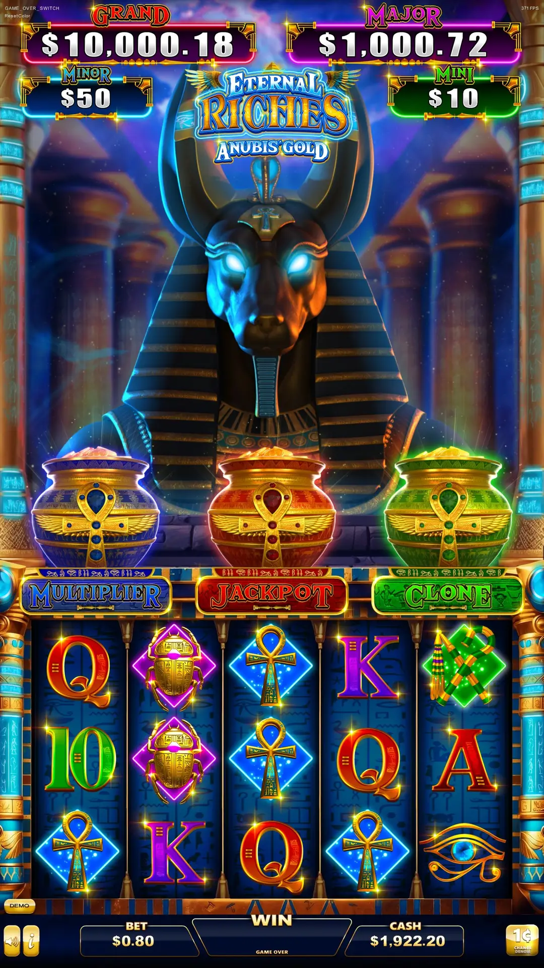 Slot machine game screen with an Egyptian Anubis statue, glowing blue eyes, and three golden spin wheels. Symbols include letters, ankhs, and scarabs. Jackpot and cash amounts are displayed at the top and bottom.