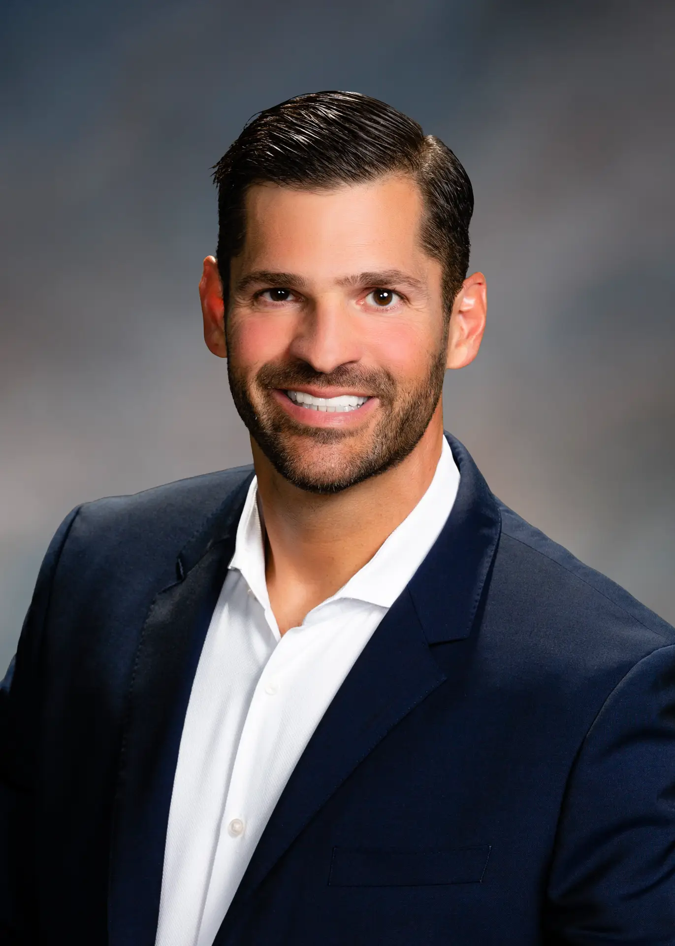 A man with short dark hair and a trimmed beard, wearing a navy suit jacket and white shirt, smiles at the camera in a professional studio portrait with a blurred gray background, exuding the confidence often seen in those pursuing successful casino gaming careers.