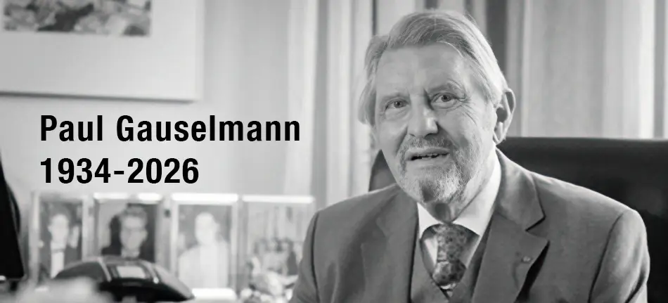 Older man with white hair and beard in a suit sits at a desk, smiling. Text beside him reads In Memoriam: Paul Gauselmann 1934-2026. Blurred family photos are visible in the background.